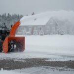 Snow blower clearing snow from a gravel driveway in front of a rural house during winter