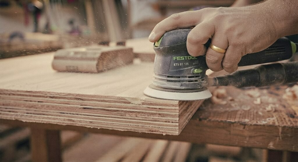 Using an orbital sander on a narrow plywood edge in a workshop.
